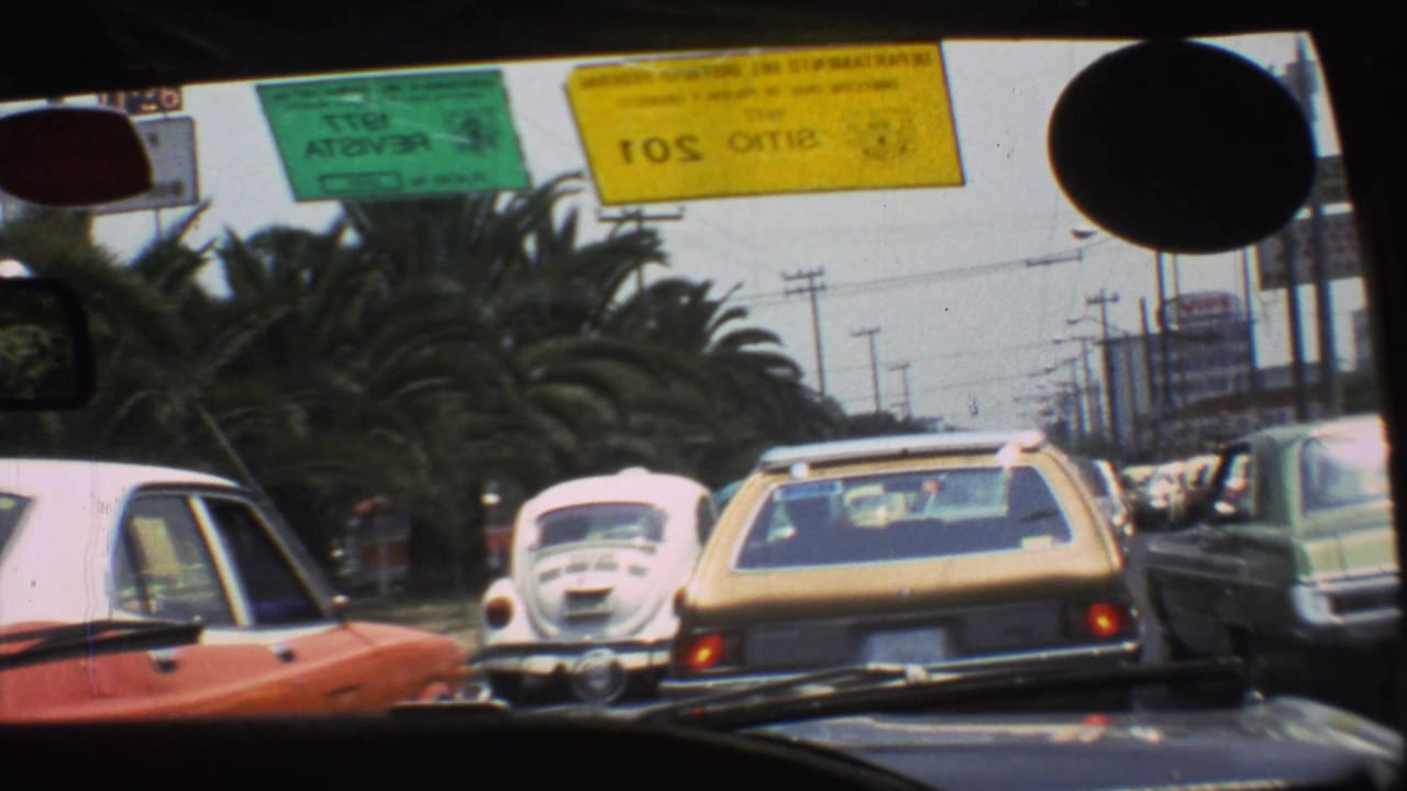 1973 Mexico City traffic through a windshield, showing a white Beetle, a yellow wagon, roadside cars, utility poles, and inspection stickers.