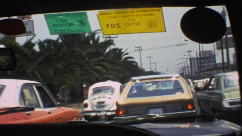 1973 Mexico City traffic through a windshield, showing a white Beetle, a yellow wagon, roadside cars, utility poles, and inspection stickers.