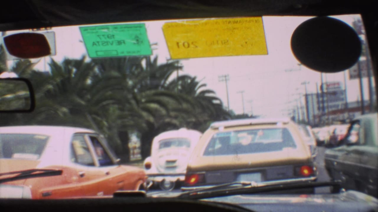 1973 Mexico City traffic viewed from inside a vehicle, with classic cars ahead, palm trees, and colored windshield stickers.