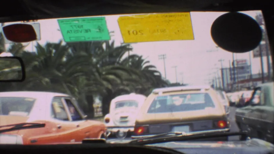 1973 Mexico City traffic viewed from inside a vehicle, with classic cars ahead, palm trees, and colored windshield stickers.