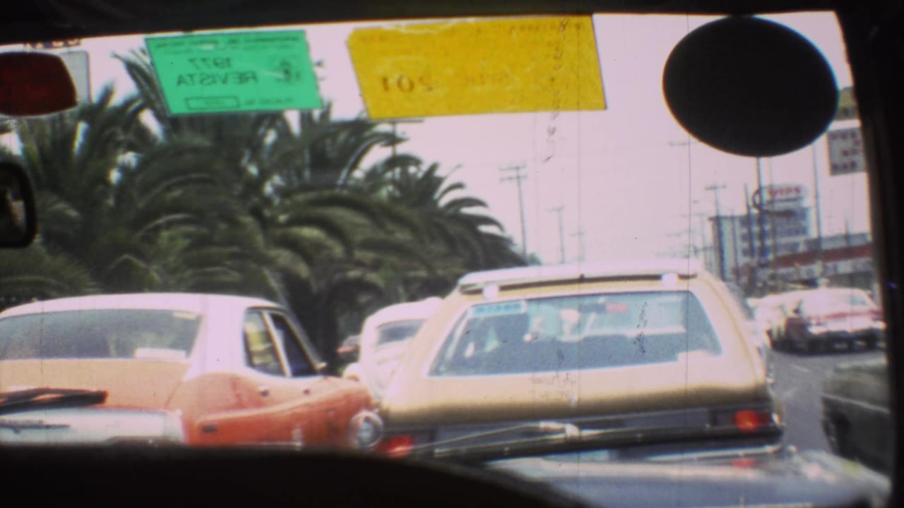 Windshield view of congested traffic with a station wagon, red car, palm trees, and road signs.