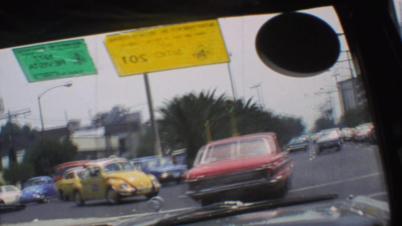 1973 Mexico City street traffic through a tilted windshield, with a red sedan, Volkswagen Beetle, palms, and multiple cars.