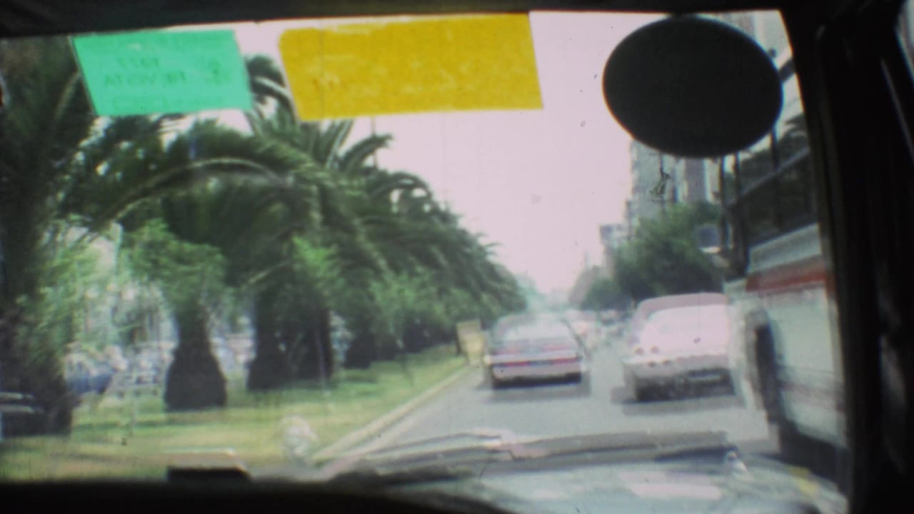 1973 Mexico City 8mm frame from inside a vehicle, looking past windshield stickers toward cars on a palm-lined street.