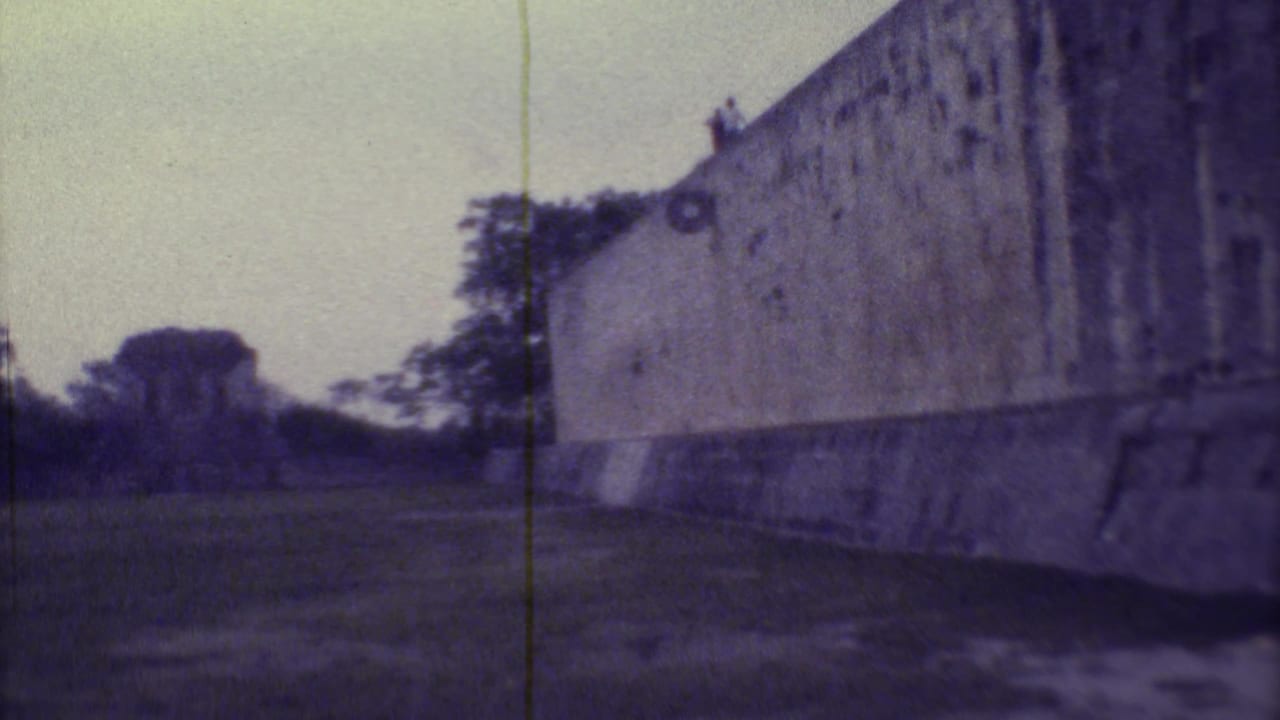 Weathered stone wall beside a grassy courtyard with trees and a distant structure.