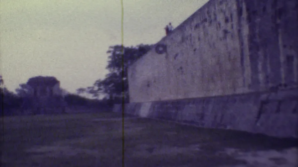 1973 Yucatan 8mm frame showing a long stone wall, open grass, trees, pale sky, and a small distant structure.