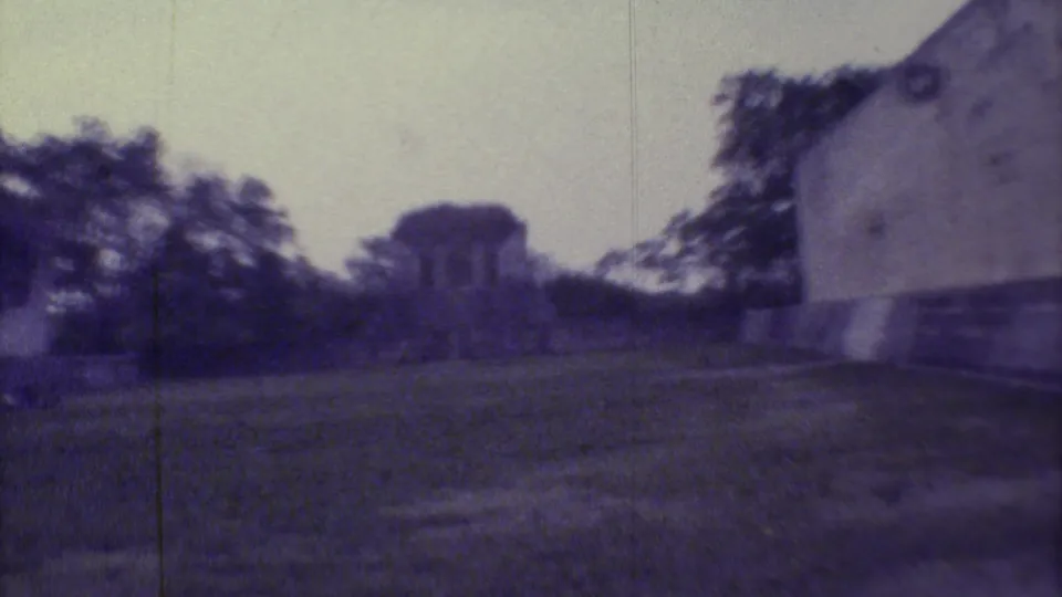 1973 Yucatan travel frame with a weathered wall at right, open grass, trees, and a small distant structure.
