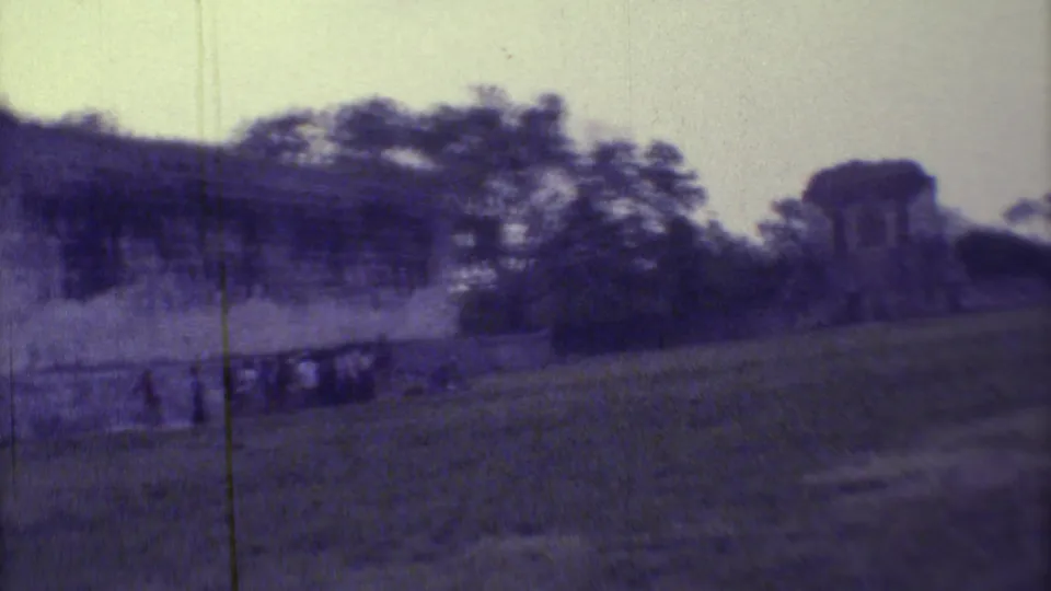 1973 Yucatan home-movie frame showing a long stone wall, distant visitors, trees, and ruin masonry.