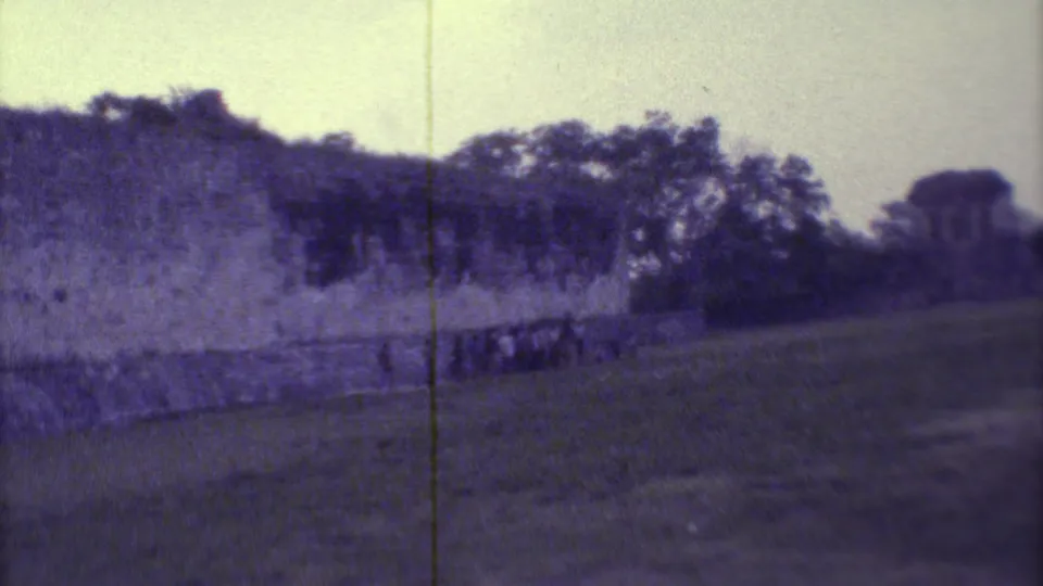 1973 8mm frame of a Yucatan ruin wall across grassy ground, with trees and a small structure in the distance.