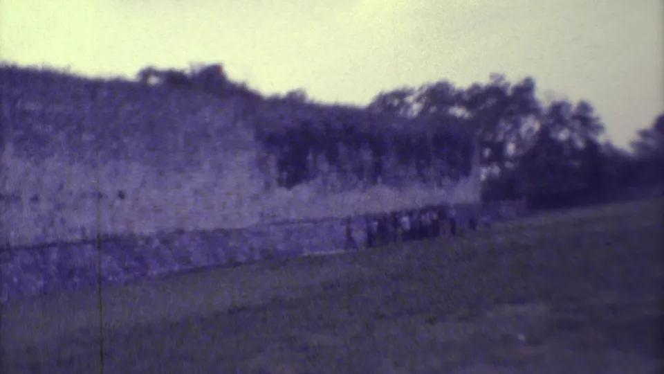 1973 Yucatan 8mm frame showing distant visitors near a long stone wall, with trees and soft, faded color.