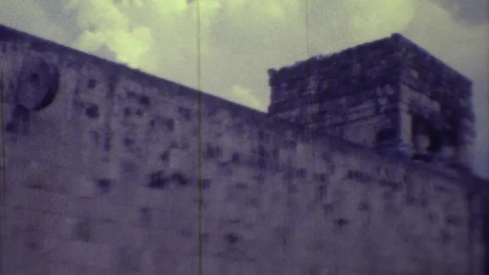 1973 Yucatan frame showing a tall stone corner structure above a long ruin wall, with grainy 8mm color and slight blur.