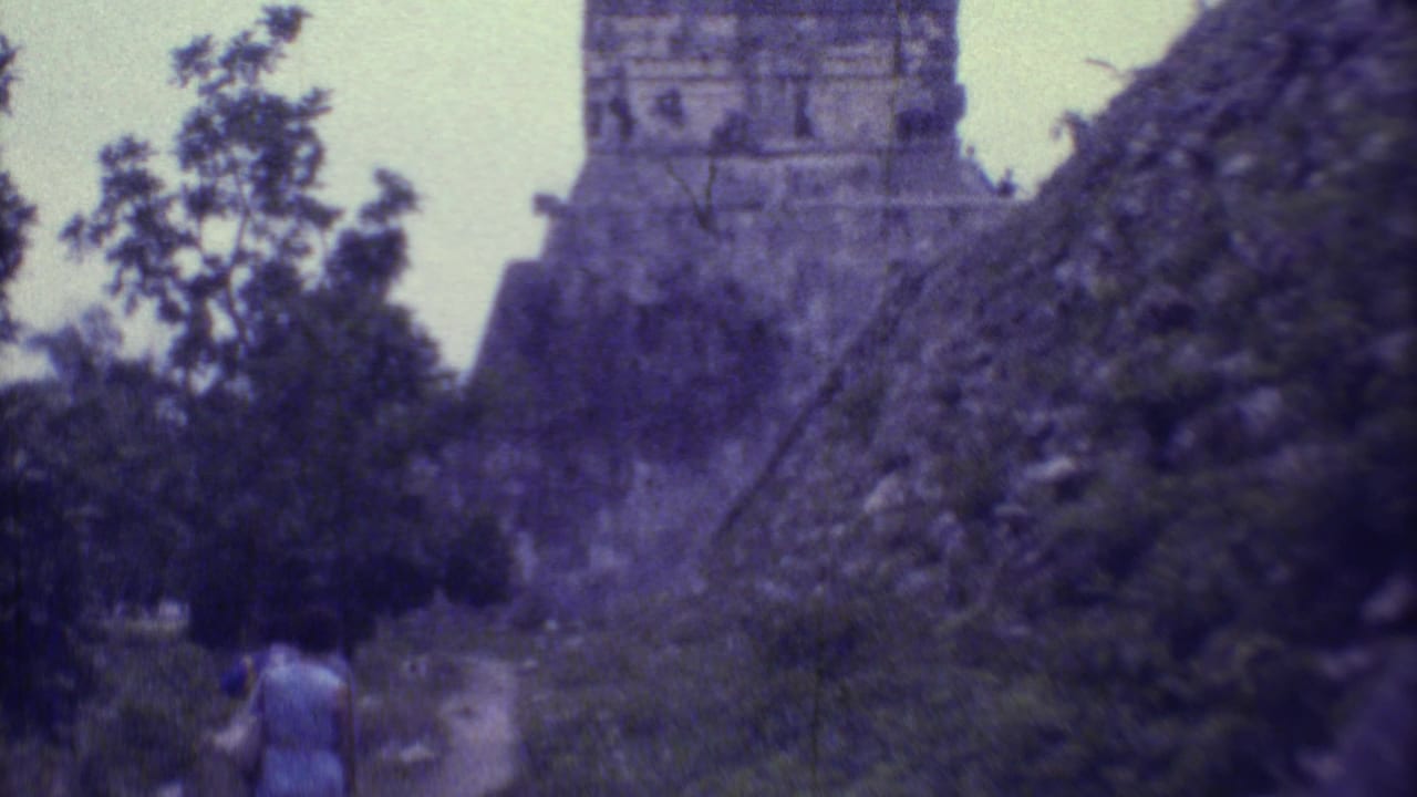 Two women walk on a narrow path toward a vine-covered stone pyramid in dense greenery.