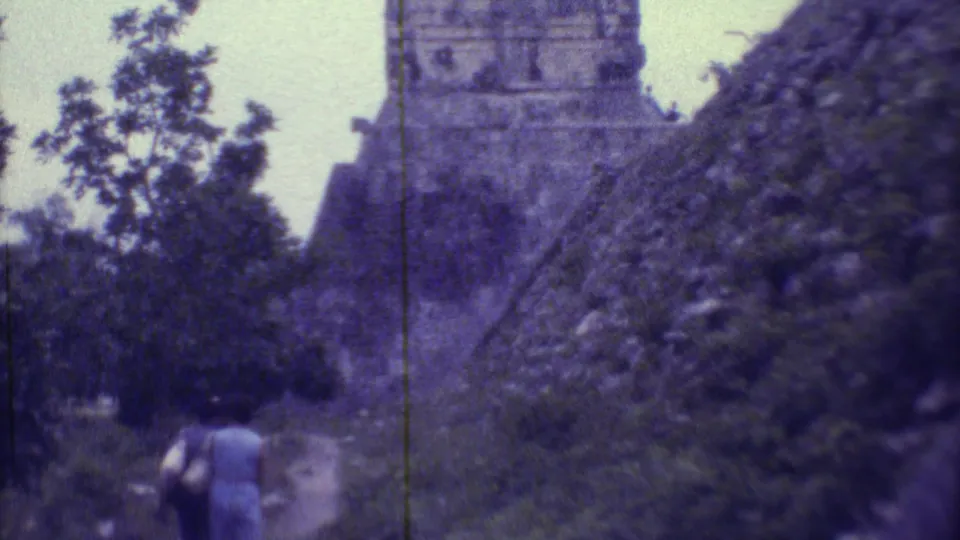 1973 Yucatan home-movie frame with a soft, purple cast, showing two women walking toward a Maya pyramid partly obscured by trees.