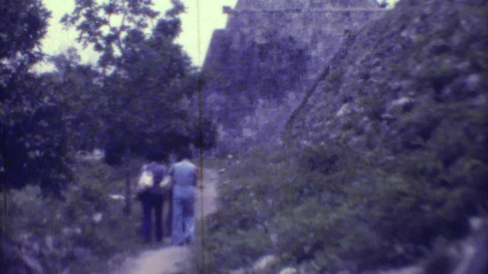 1973 8mm frame in Yucatan showing two women near dense foliage and the sloped wall of a Maya pyramid at an archaeological site.