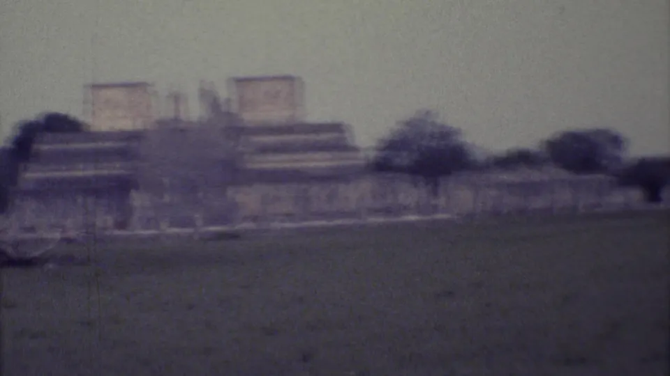 1973 8mm Yucatan travel footage shows distant Mayan stone structures beyond a grassy field, with trees and muted overcast light.