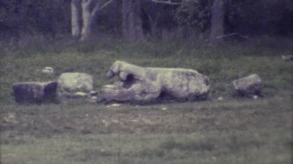 A weathered stone carving or ruin rests in grass near the jungle edge at Chichen Itza in this 1973 8mm frame.