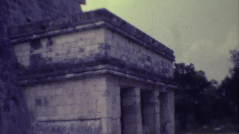 1973 8mm Yucatan frame of a stone ruin with a columned opening, dark masonry wall, trees, and pale sky.