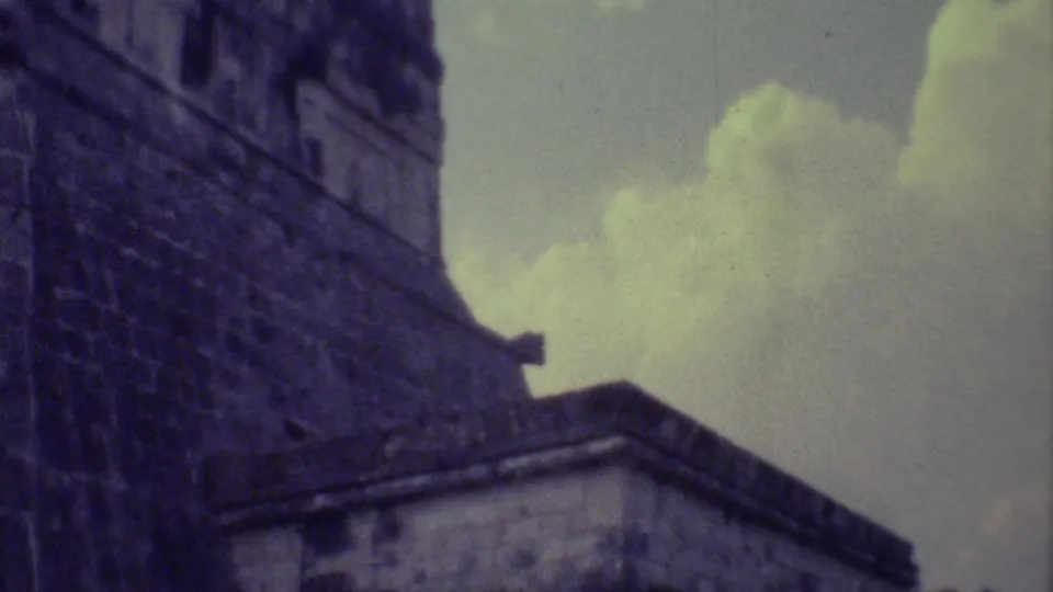 1973 Yucatan home-movie frame with a tall stone wall, lower Mayan ruin structure, dark foliage, and bright clouds.