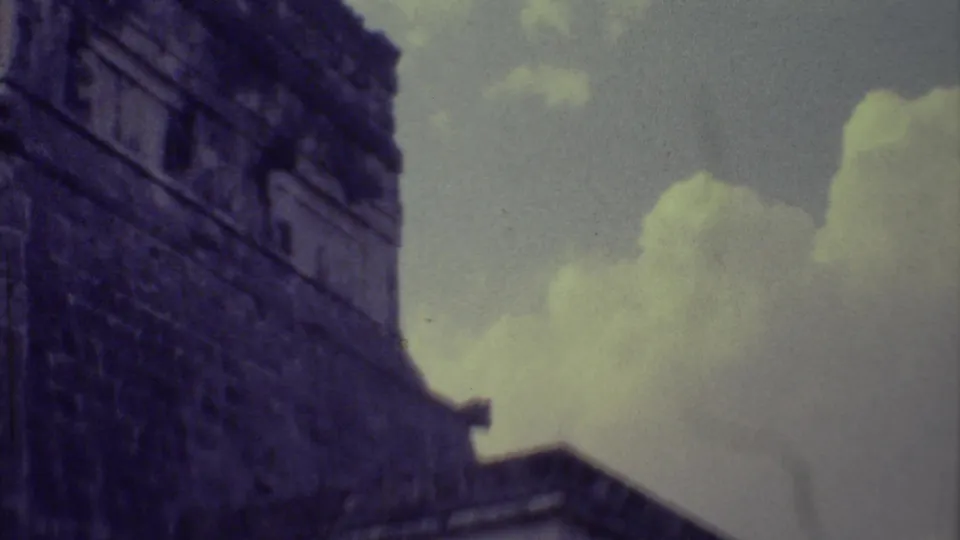 1973 8mm Yucatan footage looking upward at a dark, weathered stone ruin wall beneath pale clouds.