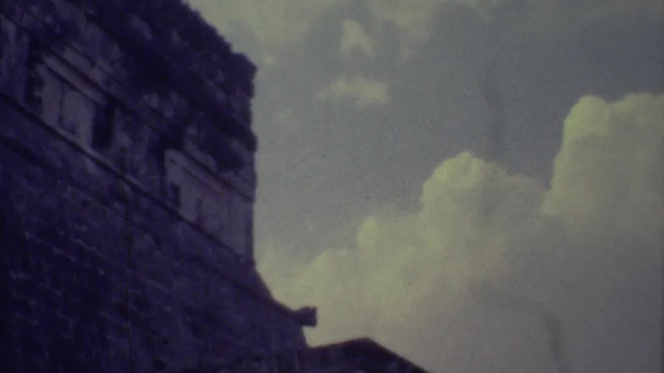 A handheld 8mm frame shows a tall Mayan ruin wall in Yucatan with large pale clouds behind it.