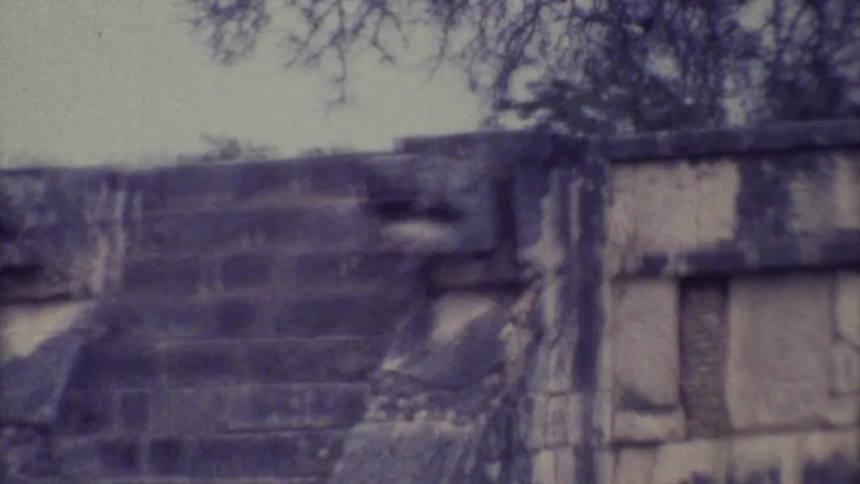 1973 8mm view of a weathered stone ruin in Yucatan, Mexico, with stepped masonry, grass, and trees in soft focus.