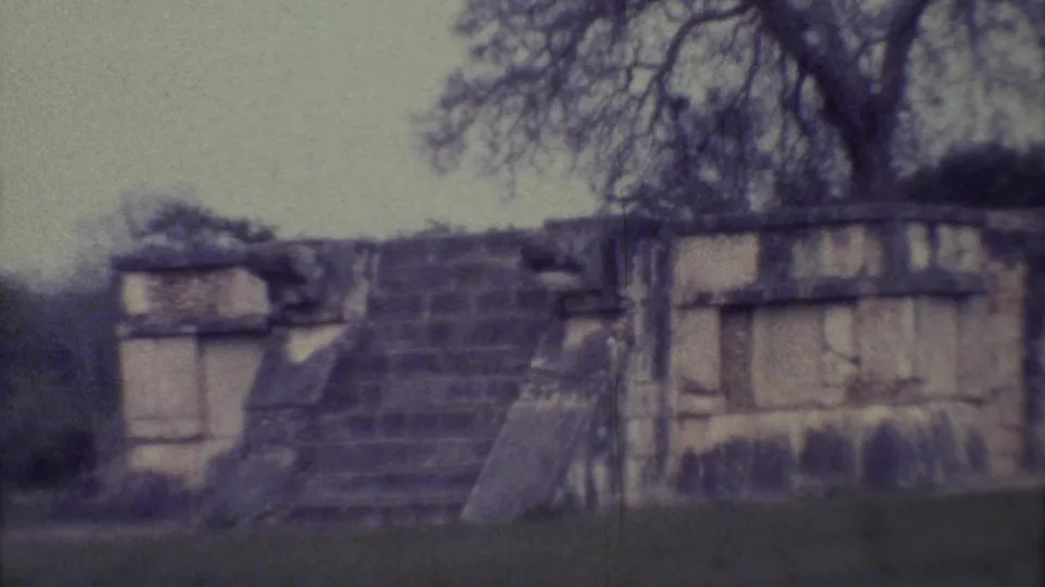 A wider 1973 Yucatan home-movie frame shows a stone temple ruin with steps, grass, shrubs, and trees behind the structure.