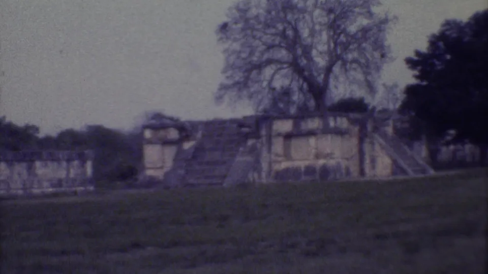 Wide 8mm frame of a Yucatan stone ruin in 1973, seen across a grassy area with large trees and soft, faded film color.