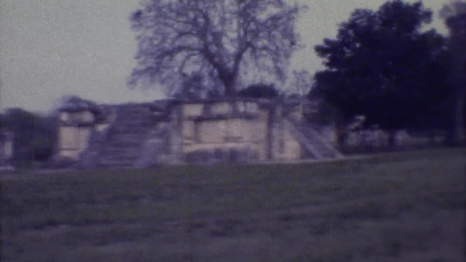 1973 Yucatan home-movie frame showing weathered stone ruins with steps, grassy foreground, and trees in soft faded color.