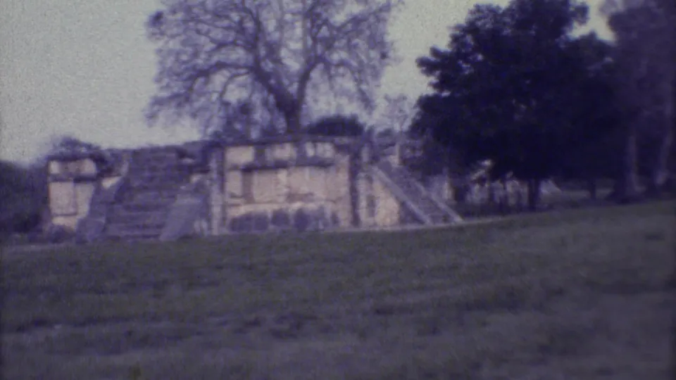 Soft 8mm frame from Yucatan showing stepped ruins, a grassy approach, and trees during a handheld pan.