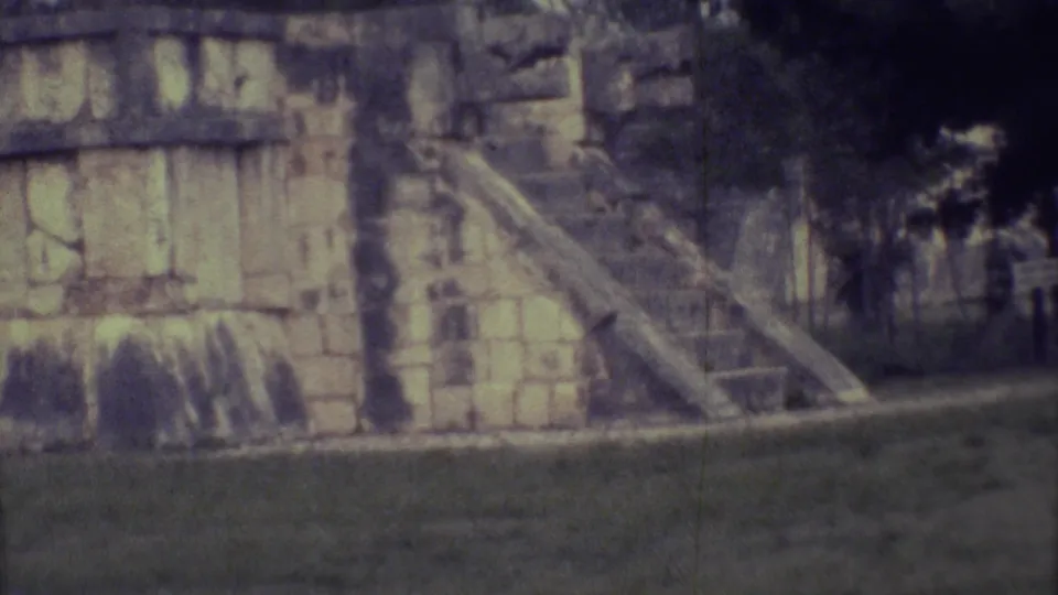 1973 Yucatan 8mm frame holding on weathered stonework, an exterior stairway, grass, and shadowed trees behind the structure.