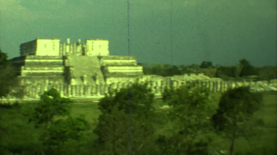 Distant 1973 8mm view of a stepped Mayan temple structure at Chichen Itza with trees and grass in the foreground.