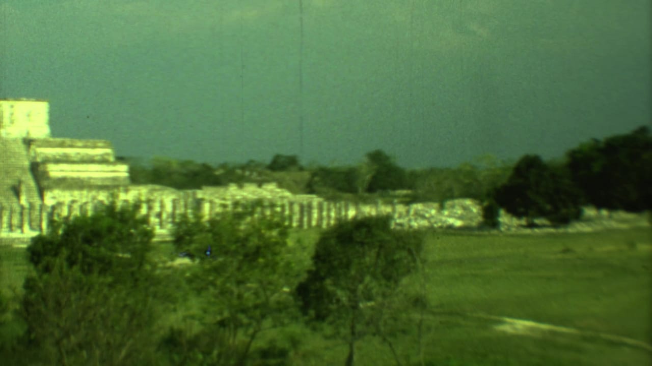 1973 8mm view of Mayan ruins in Yucatan, Mexico seen from a distance across grass, trees and a hazy sky.