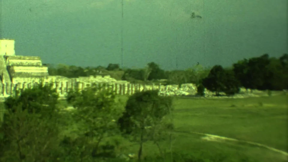 Distant Yucatan Mayan ruins on 8mm film, with a stepped stone form, low masonry, grass and trees visible.