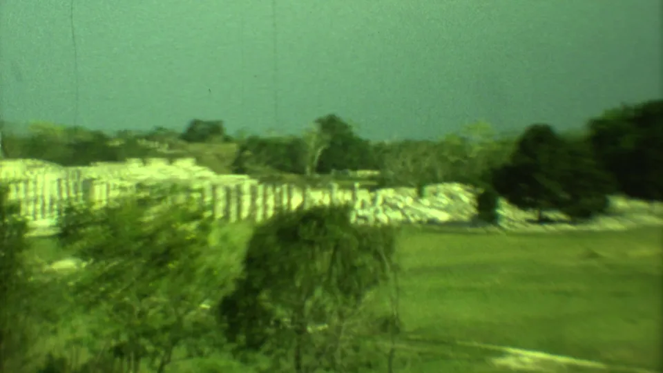 1973 Yucatan home-movie frame showing low Mayan ruins partly hidden by trees across an open grassy area.