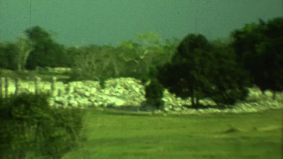 Handheld 8mm frame from Yucatan showing open grass, dark trees, and pale ruin stonework under a hazy sky.