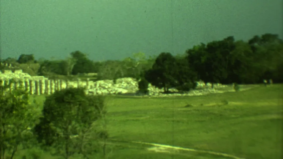 Yucatan 8mm travel frame showing grass, trees, pale ruins, and tiny distant visitors near the stonework.