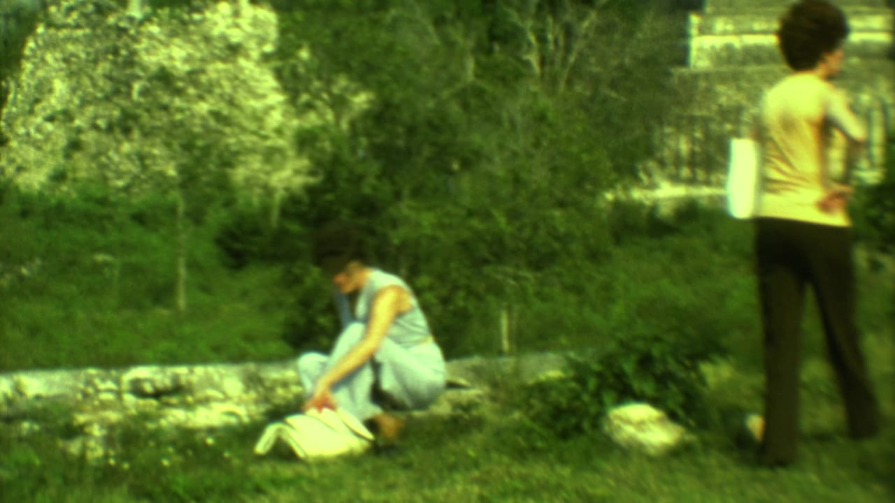 Two women near a low stone wall with grass and ruins in the background.
