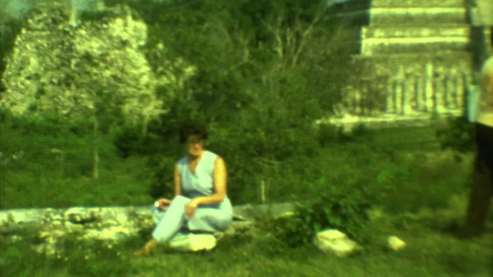 A woman in a light blue sleeveless dress sits on a low wall at a Yucatan ruin site, with another visitor partly visible nearby.