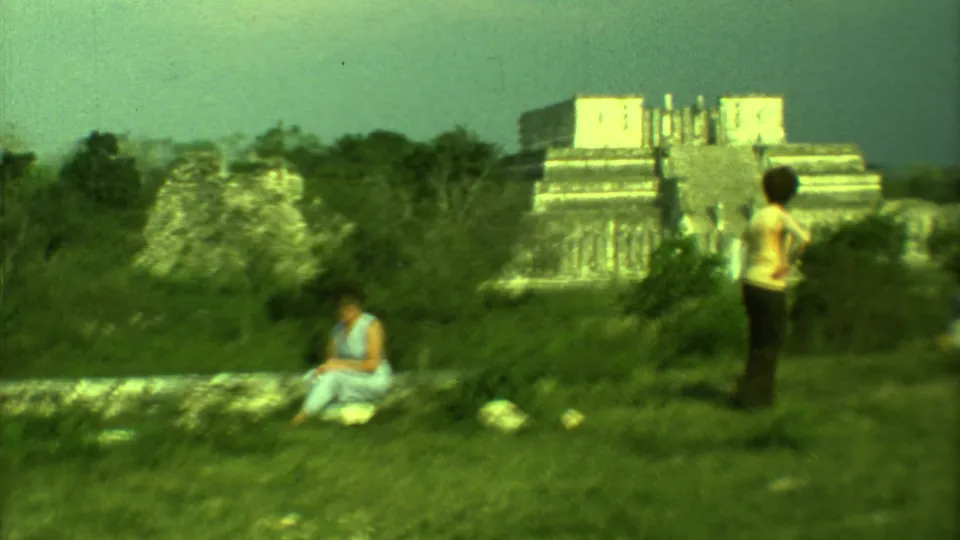 Wide 1973 Yucatan view of two women at a grassy Mayan ruin area with a stepped stone building in the distance.