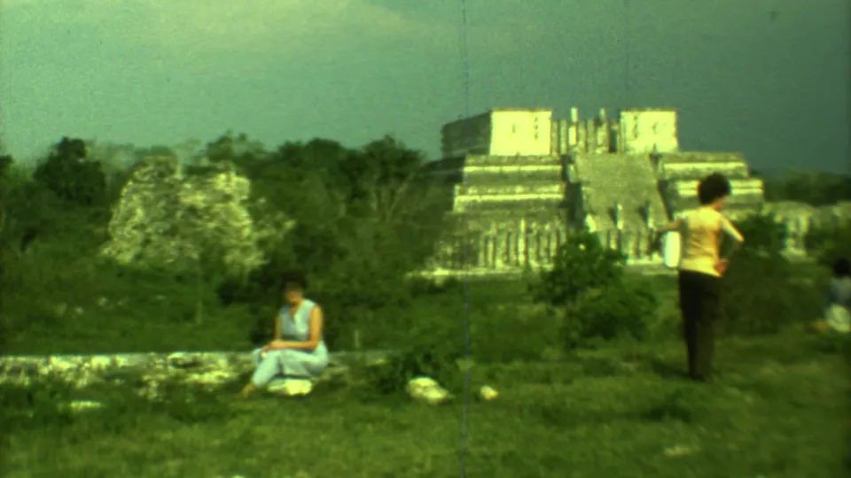 Yucatan home-movie frame showing a seated woman and a standing woman in front of Mayan stone ruins and greenery.