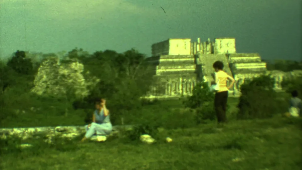 1973 Yucatan travel footage frame with a visitor standing in the foreground and a stepped archaeological structure in the distance.