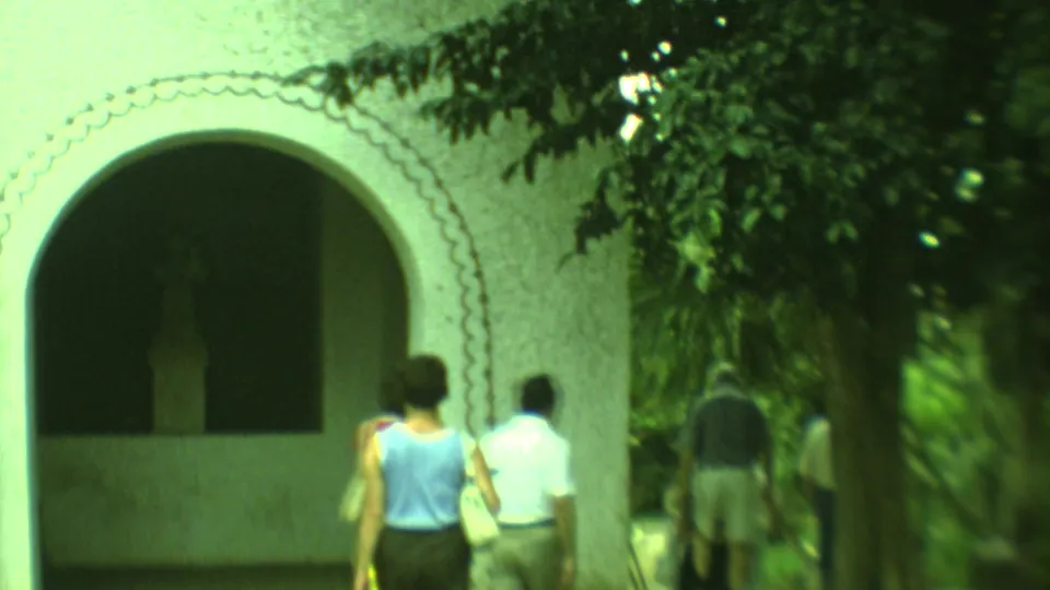 Soft 8mm frame from Yucatan showing two adults walking near a white arched church exterior and shaded foliage.