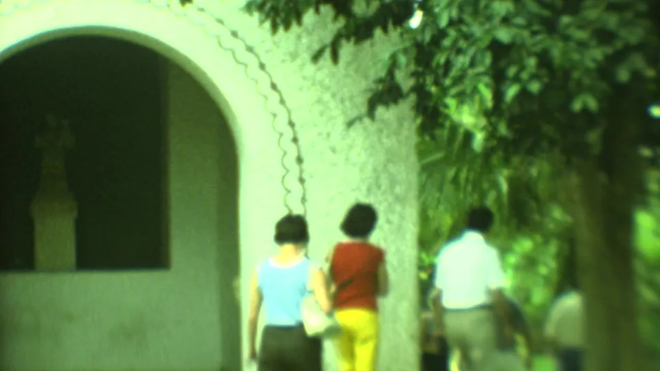 1973 Yucatan home-movie frame showing adults walking away beside a white stucco church arch and dense greenery.
