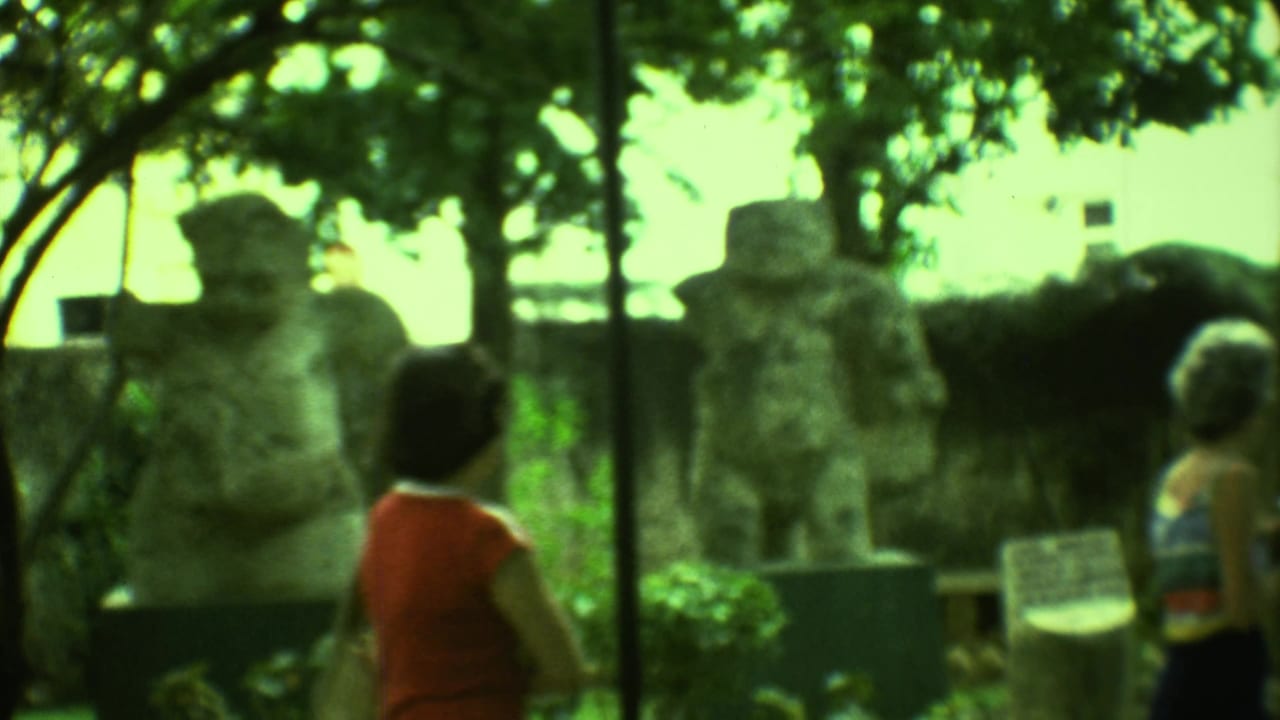 A visitor in a red top faces carved stone figures under trees.