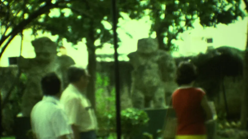 1973 Yucatan 8mm frame with tourists in summer clothing walking past large carved stone figures and trees.