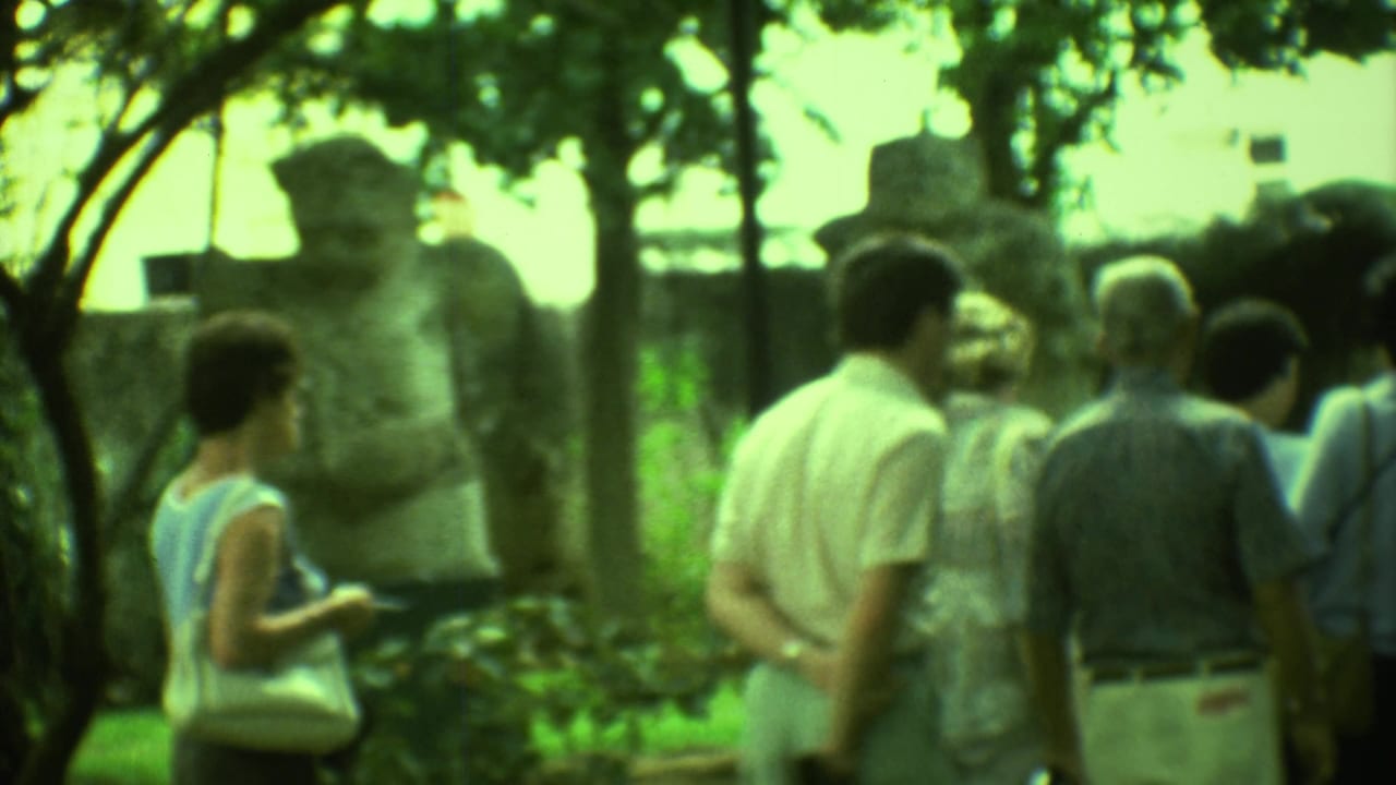 Visitors stand near a weathered stone figure under trees at an archaeological site.