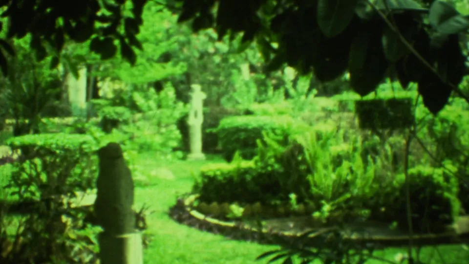 1973 Yucatan garden cutaway with a curved planting border, dense tropical greenery, lawn, and a pale stone figure under shaded leaves.