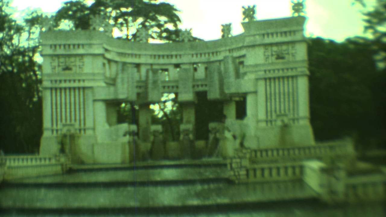 Carved stonework monument beside a low building and roadside landscaping.