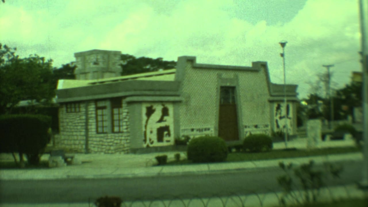 1973 Yucatan 8mm frame showing a low stone-faced building with bold mural panels, clipped shrubs, a road, and utility poles.