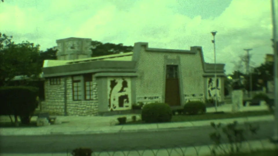1973 Yucatan 8mm frame showing a low stone-faced building with bold mural panels, clipped shrubs, a road, and utility poles.
