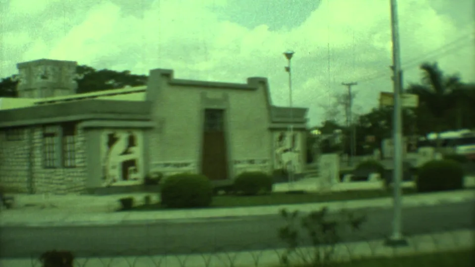1973 Yucatan 8mm frame with a roadside view of the mural-fronted low building, a lamp post, clipped shrubs, and cloudy sky.